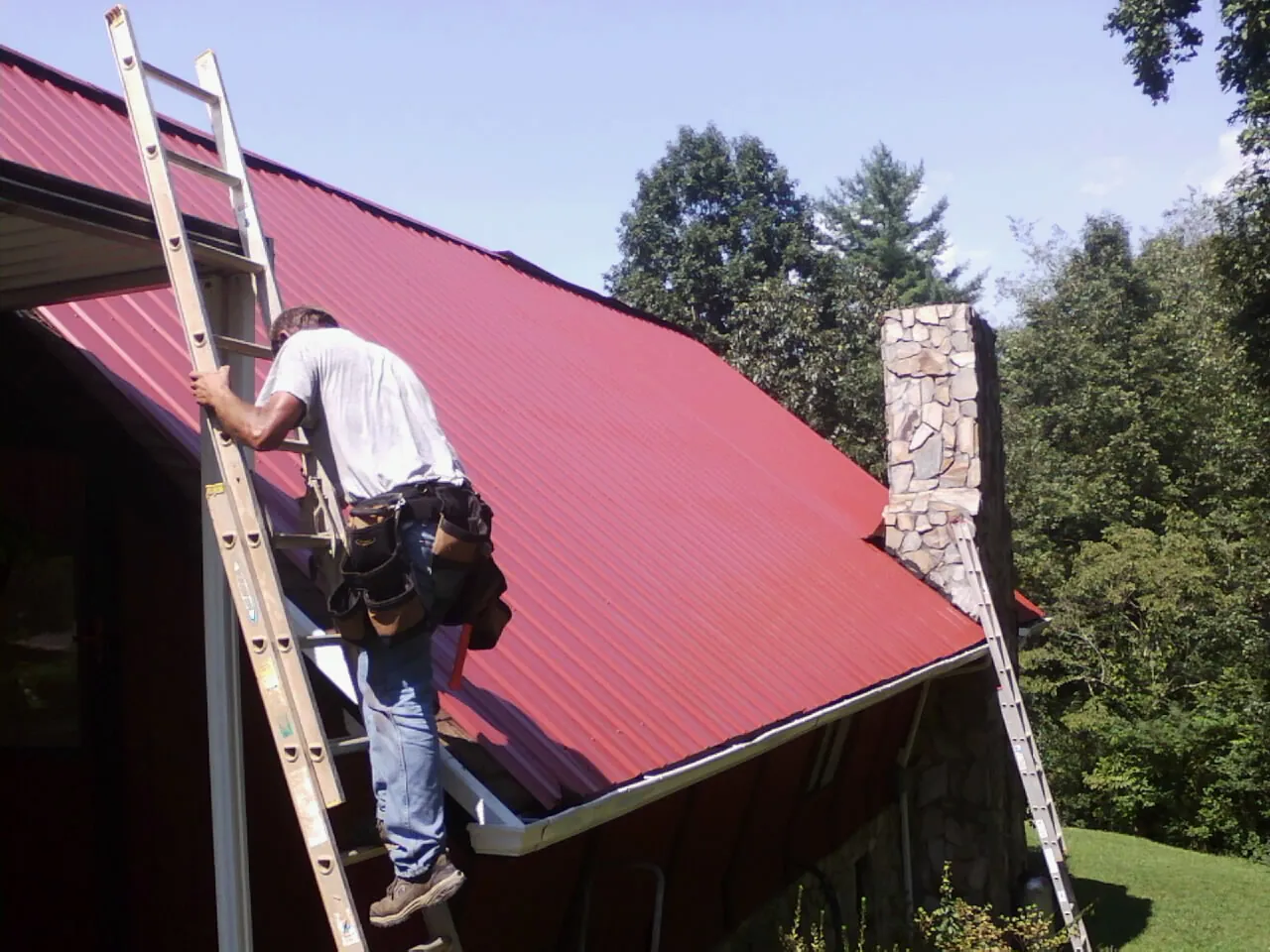 Roofer on a ladder beside a red metal roof and stone chimney — Soffit & Fascia Repair by Roofing Brothers in Plainsboro